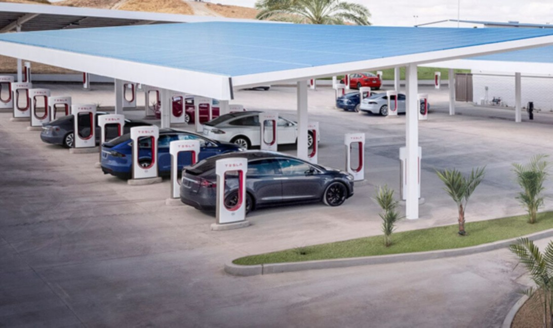 Ilmakuva Tesla Supercharger -latausasemasta, jossa useita Tesla-autoja latauksessa sinisen katoksen alla.